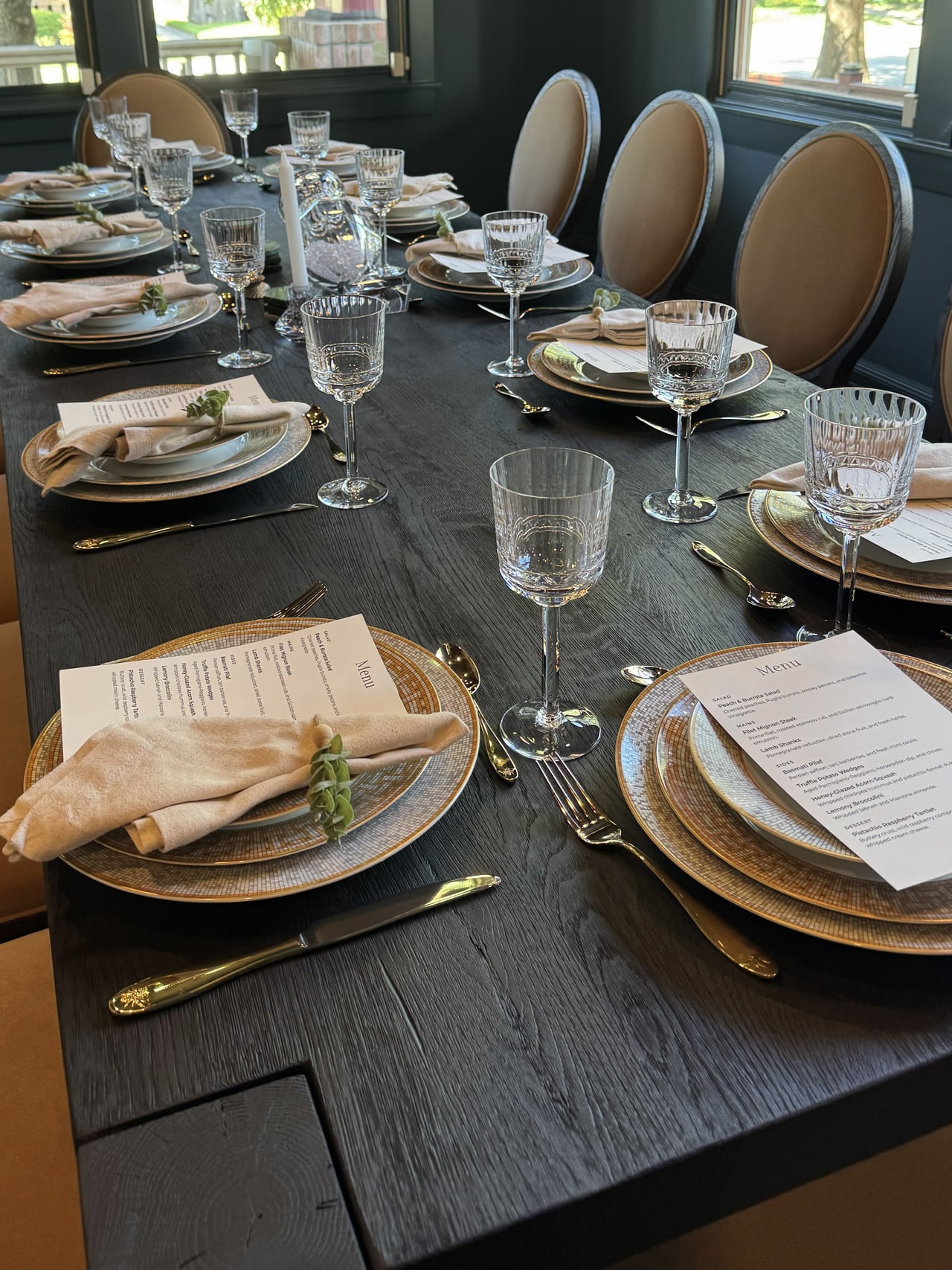 A dining table set with printed menus, gold-rimmed plates, folded napkins and glassware for the Houston housewarming dinner.