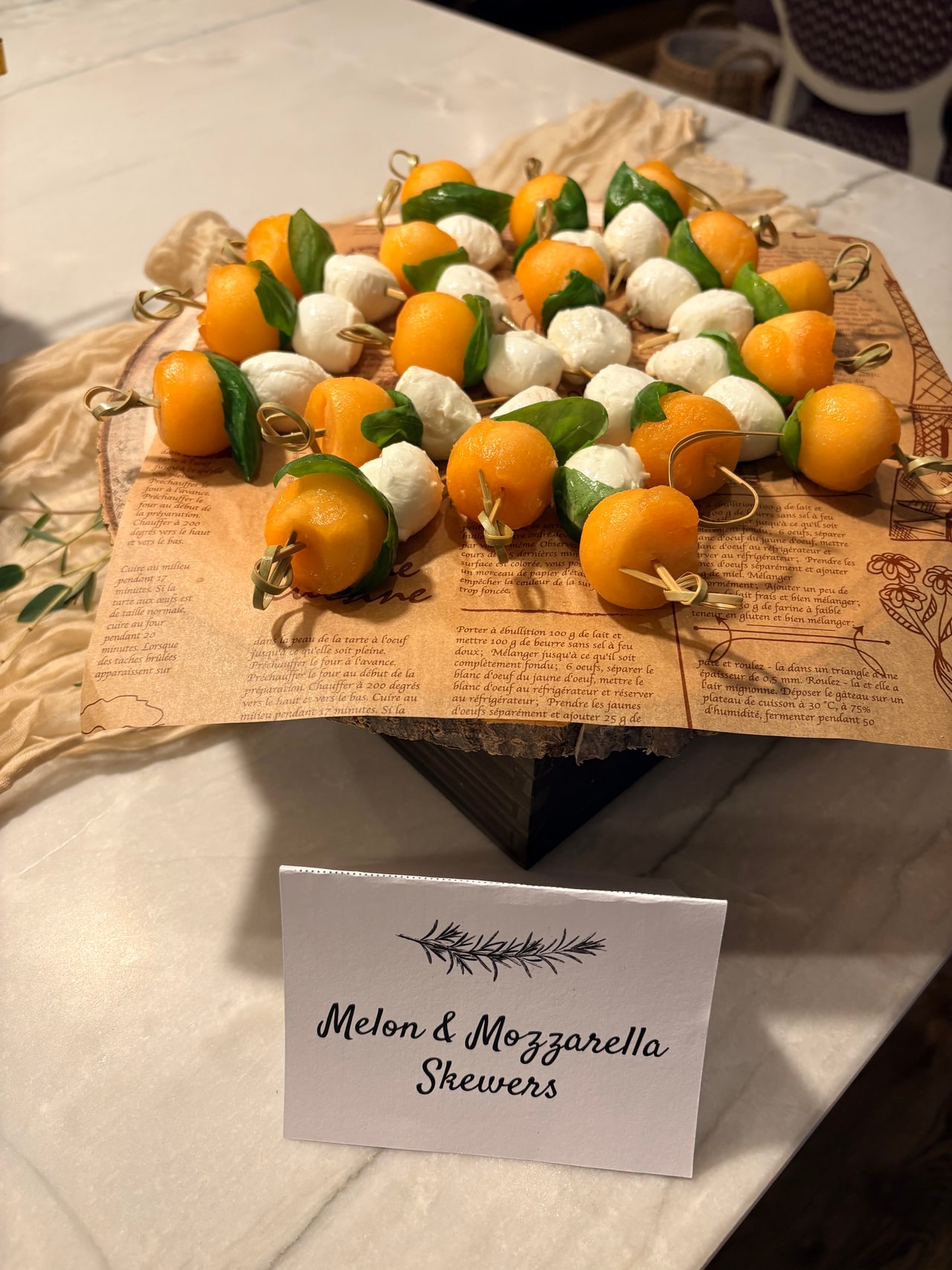 Melon balls, mozzarella pearls and basil leaves on skewers, displayed beside a printed dish label.