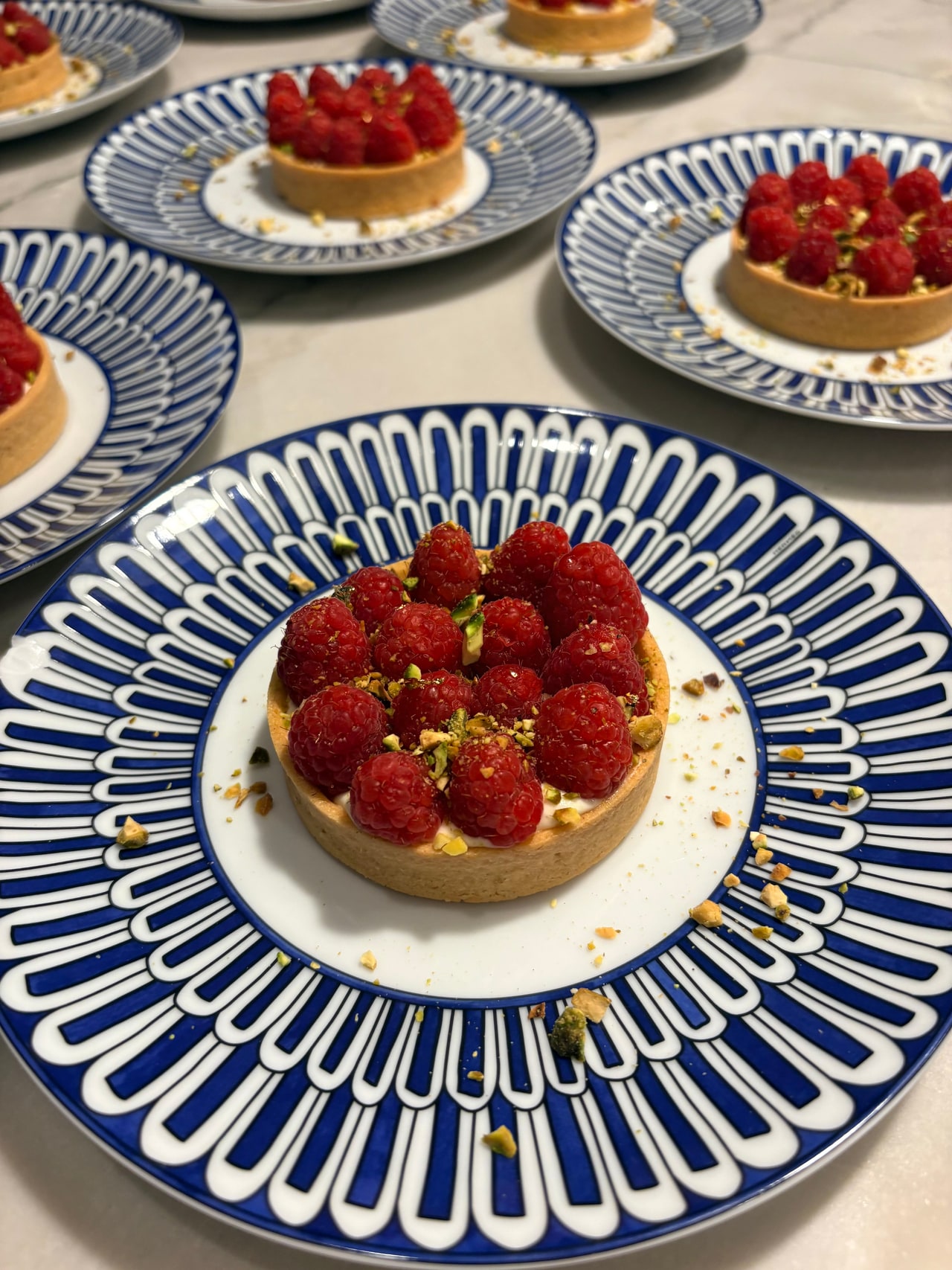 Individual raspberry tartlets finished with pistachio crumbs and gold leaf on blue-and-white dessert plates.