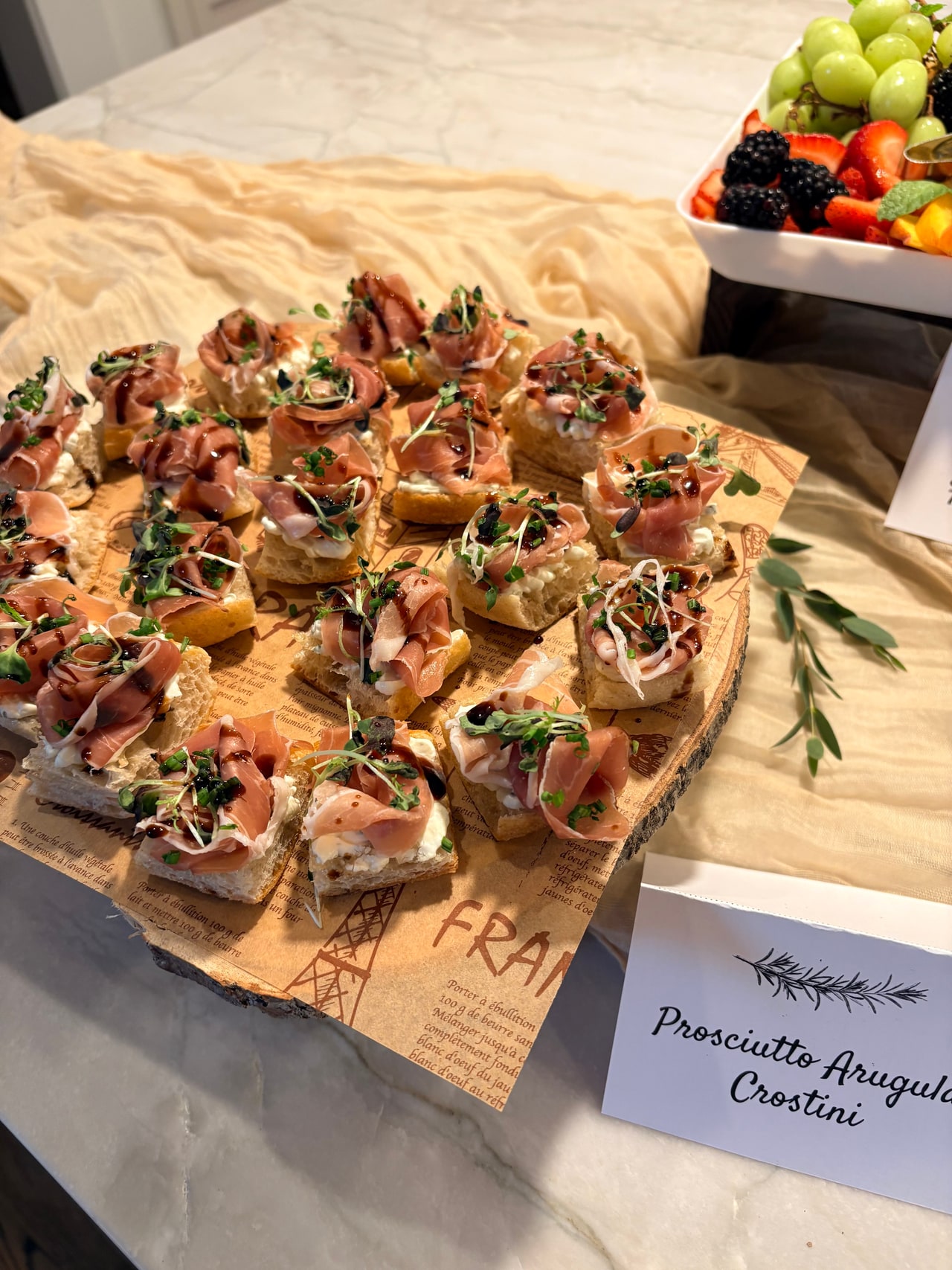 Prosciutto and arugula crostini arranged on a wooden board beside their printed menu label.